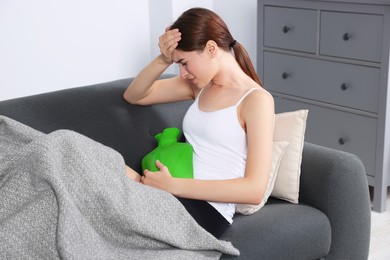 Young woman using hot water bottle to relieve cystitis pain on sofa at home