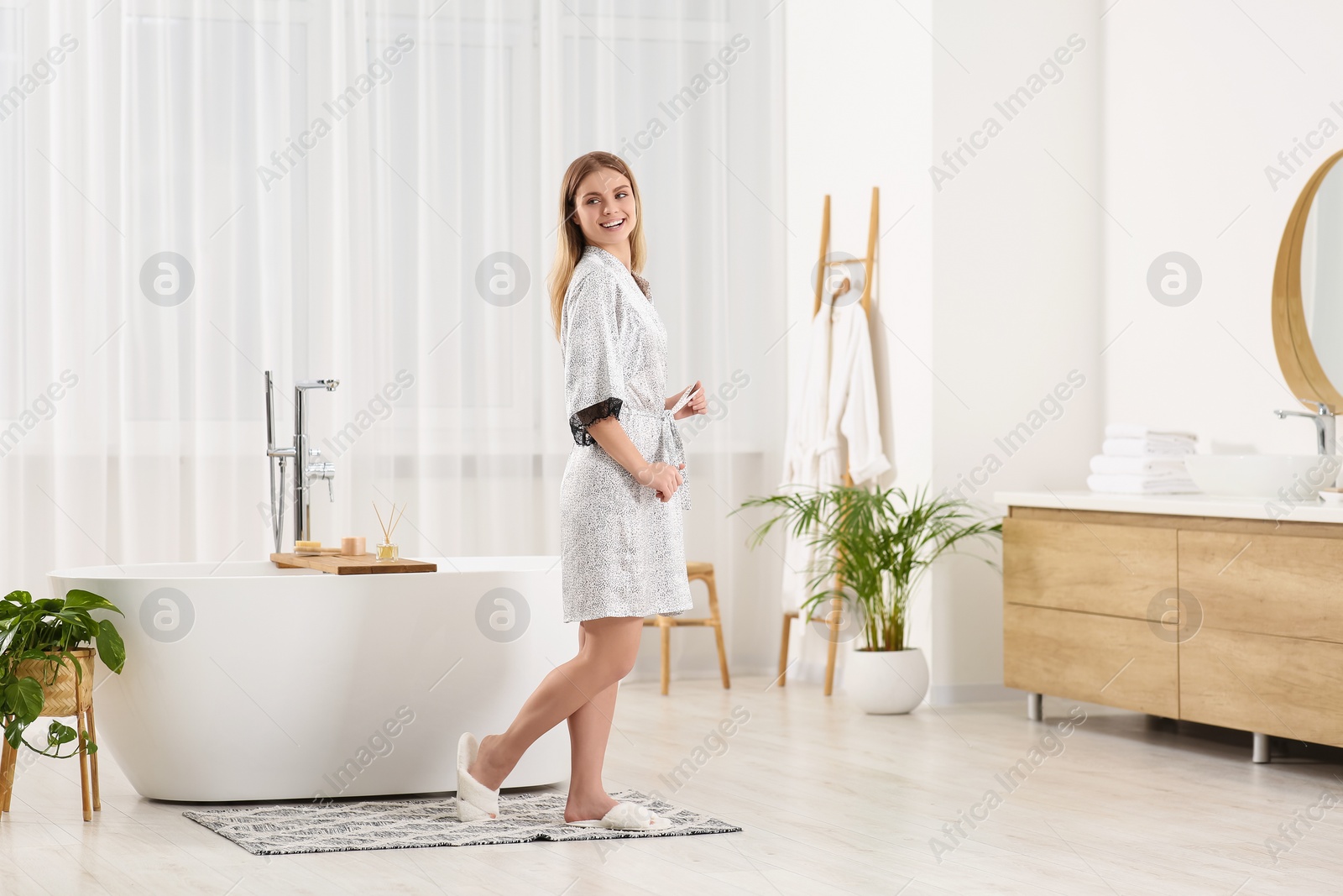 Photo of Beautiful woman wearing stylish robe near tub in bathroom