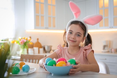 Photo of Cute little girl in bunny ears headband painting Easter eggs at table indoors