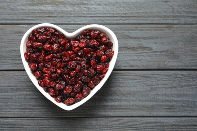 Photo of Heart shaped bowl with cranberries on wooden background, top view with space for text. Dried fruit as healthy snack