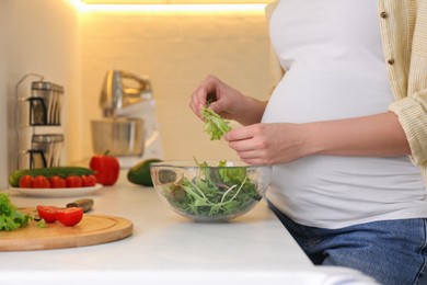 Photo of Young pregnant woman preparing vegetable salad at table in kitchen, closeup. Healthy eating