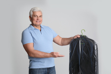 Senior man holding hanger with jacket in plastic bag on light grey background. Dry-cleaning service