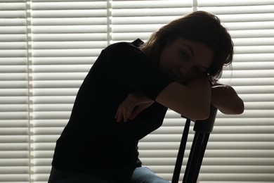 Photo of Silhouette of sad young woman near closed blinds indoors