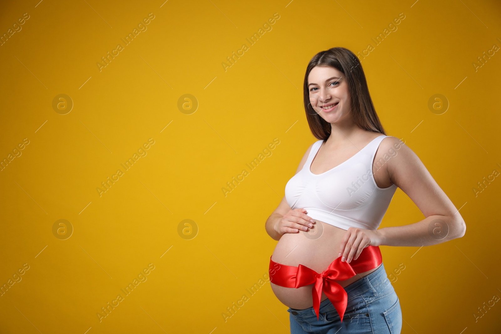 Photo of Young pregnant woman with red bow on her belly against yellow background, space for text. Time to give birth
