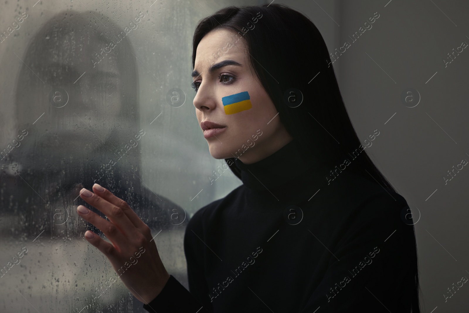 Image of Upset young woman with picture of Ukrainian flag on cheek near window at home. Stop war in Ukraine