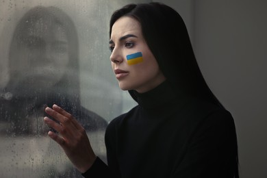 Upset young woman with picture of Ukrainian flag on cheek near window at home. Stop war in Ukraine