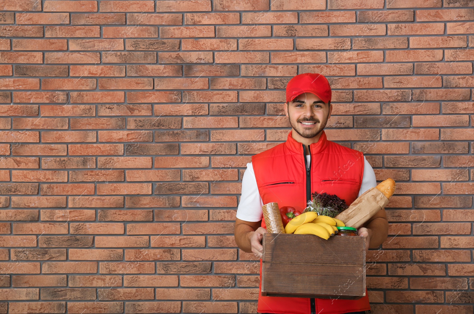 Photo of Food delivery courier holding wooden crate with products near brick wall. Space for text