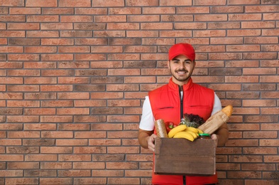 Food delivery courier holding wooden crate with products near brick wall. Space for text