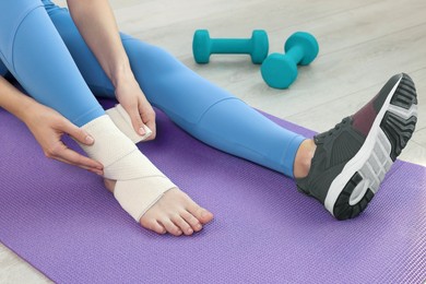 Photo of Woman wrapping foot in medical bandage on yoga mat indoors, closeup