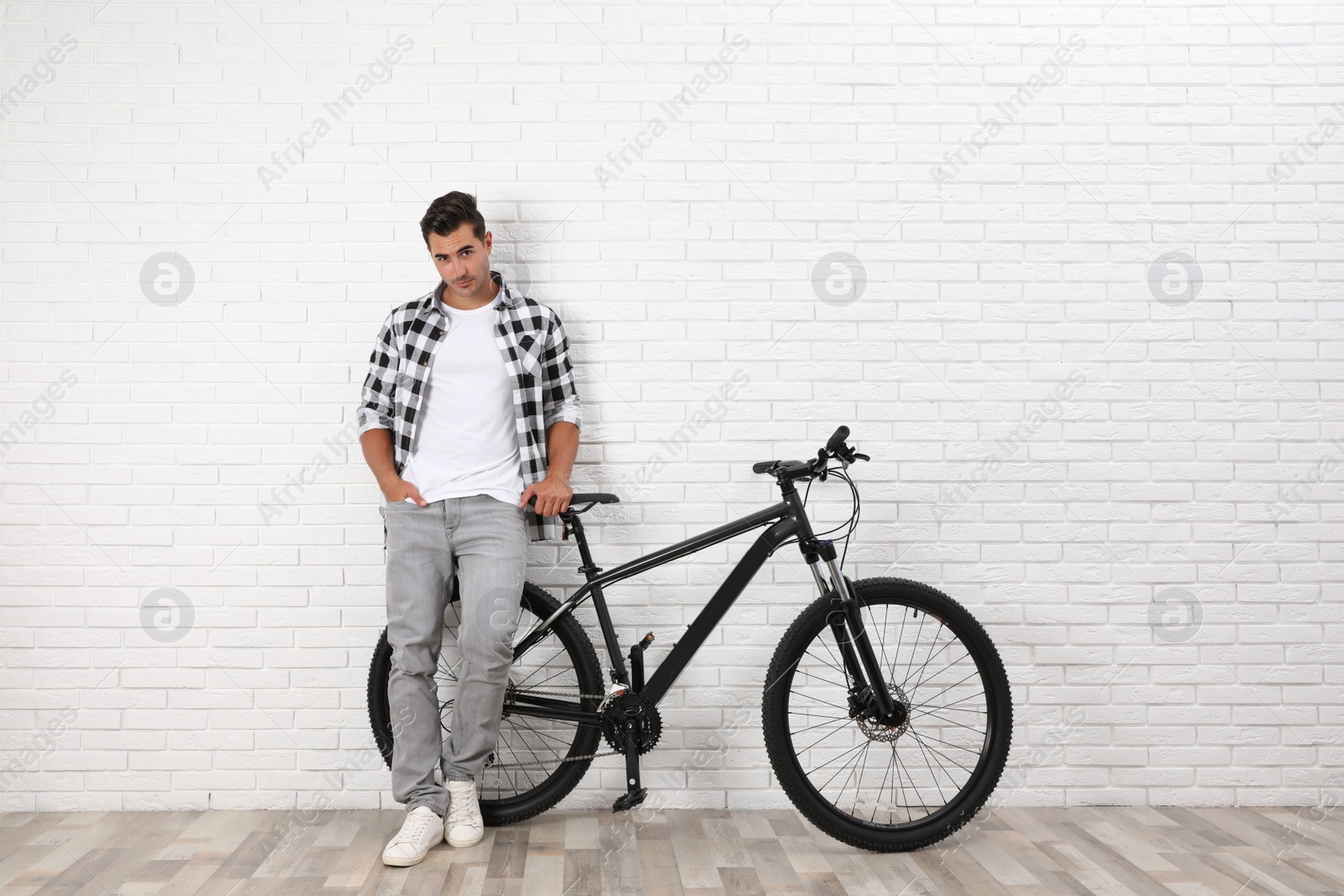 Photo of Handsome young man with modern bicycle near white brick wall indoors