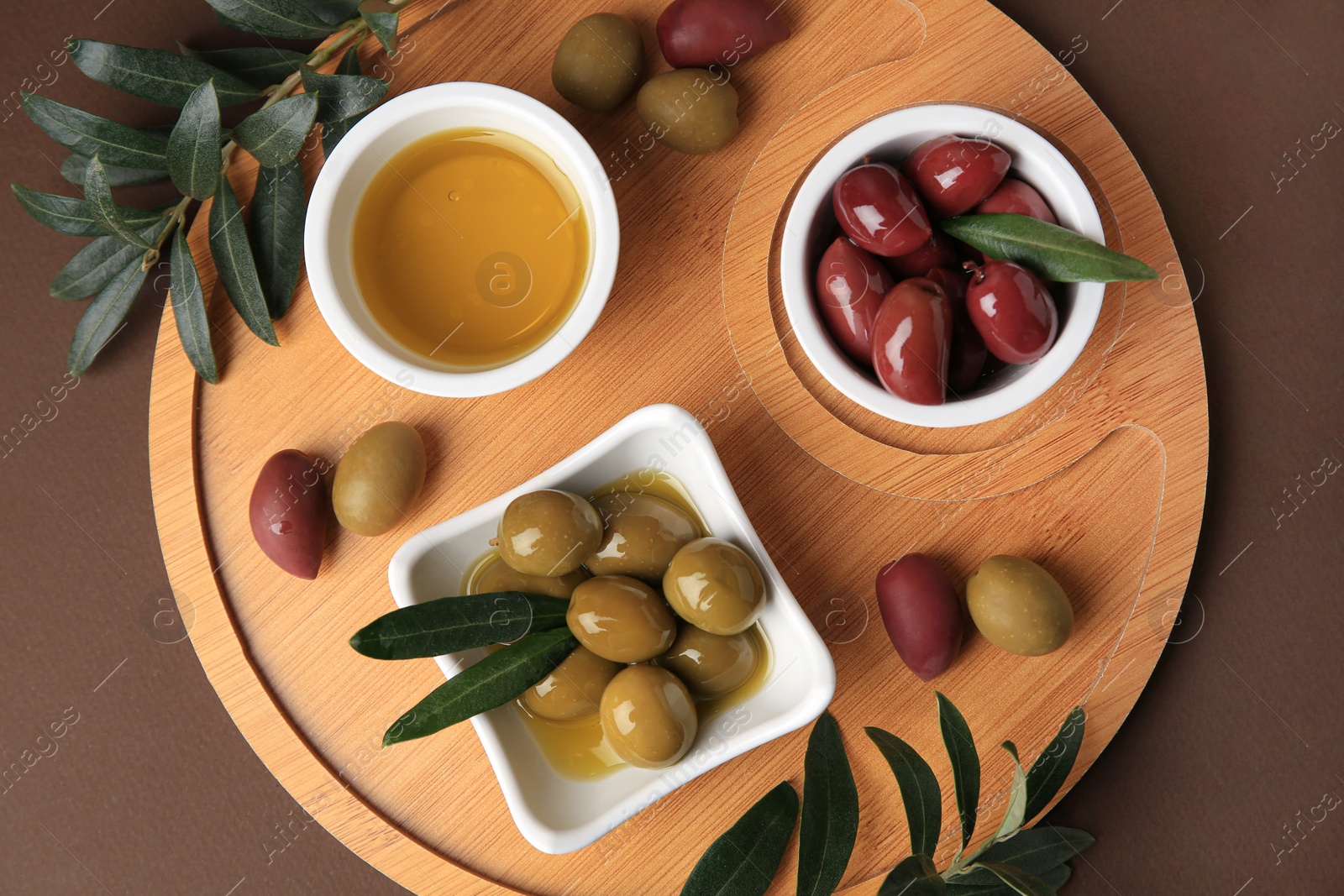 Photo of Bowl of oil, olives and tree twigs on brown table, top view