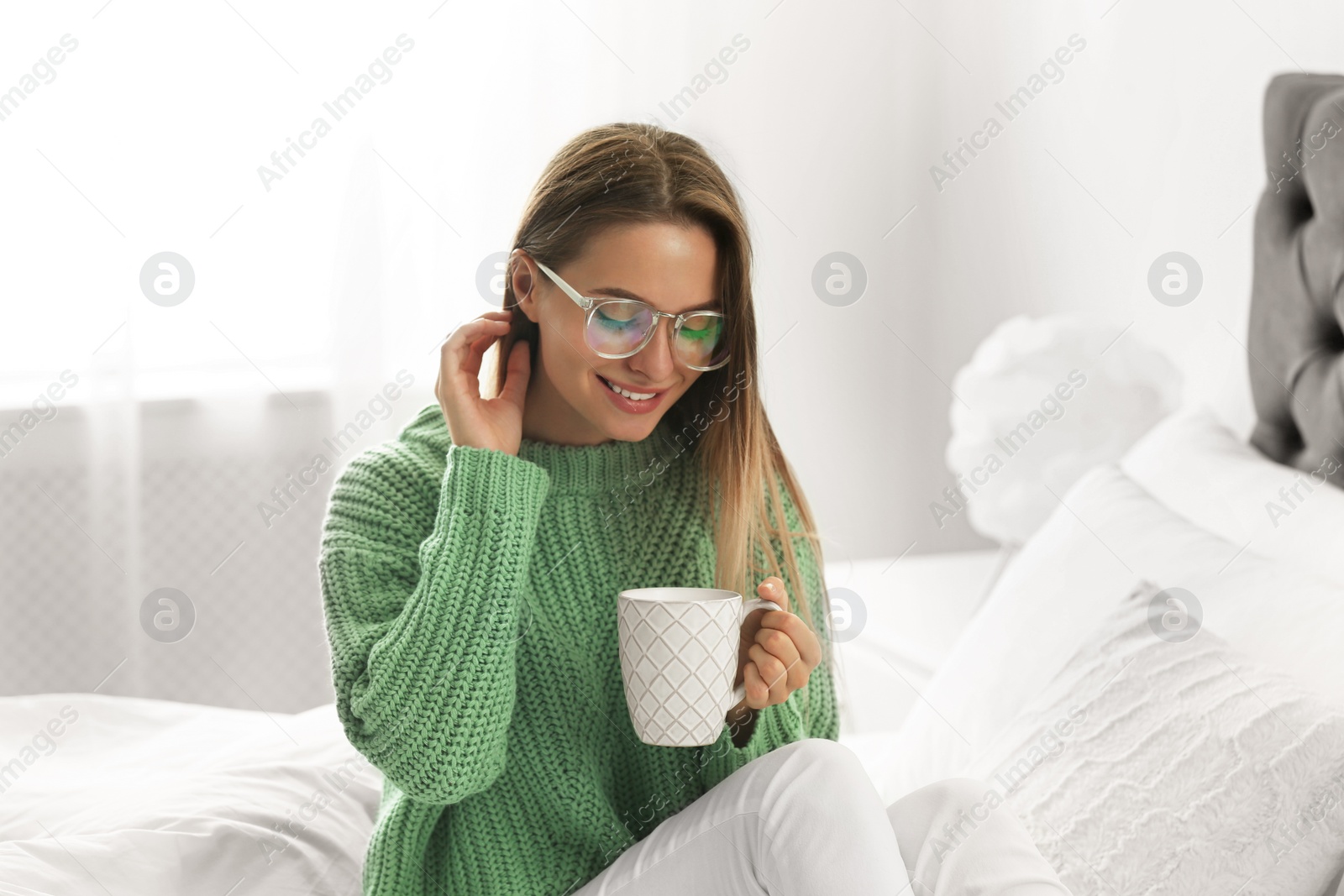 Photo of Young woman in warm sweater with hot drink on bed at home