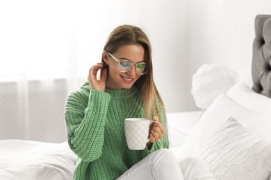 Photo of Young woman in warm sweater with hot drink on bed at home