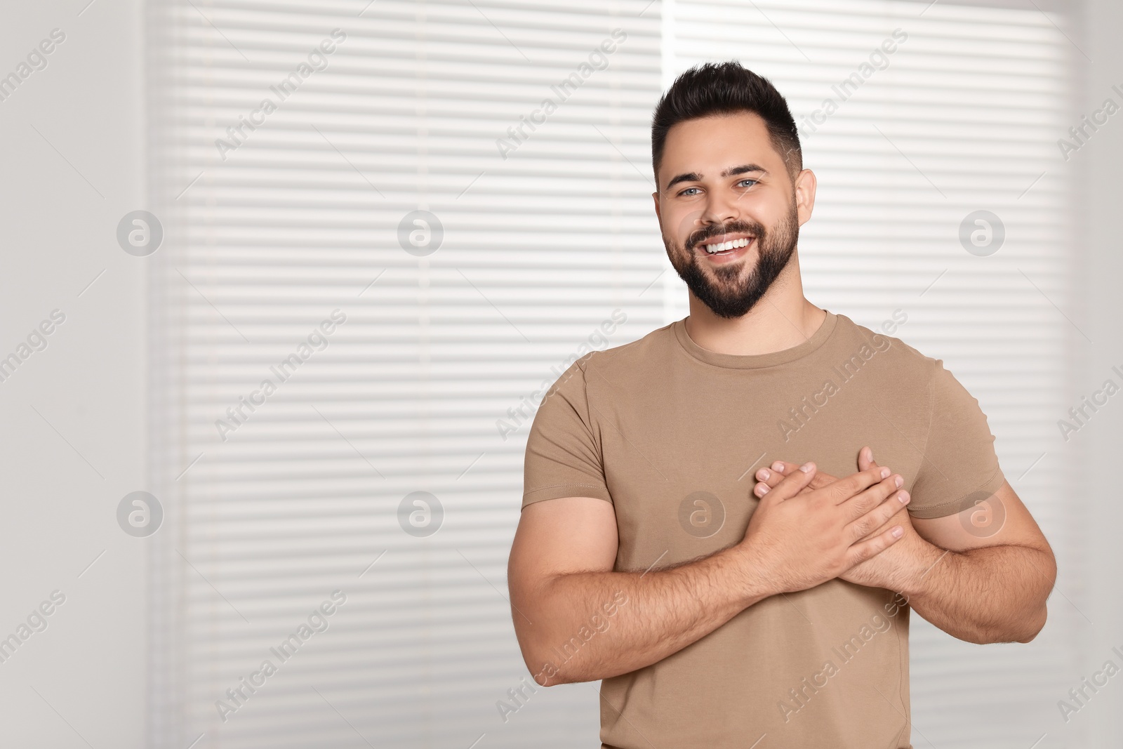 Photo of Thank you gesture. Happy grateful man holding hands near heart indoors, space for text