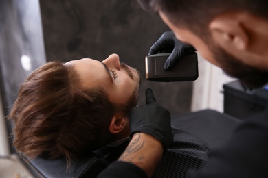 Photo of Professional hairdresser shaving client with electric razor in barbershop
