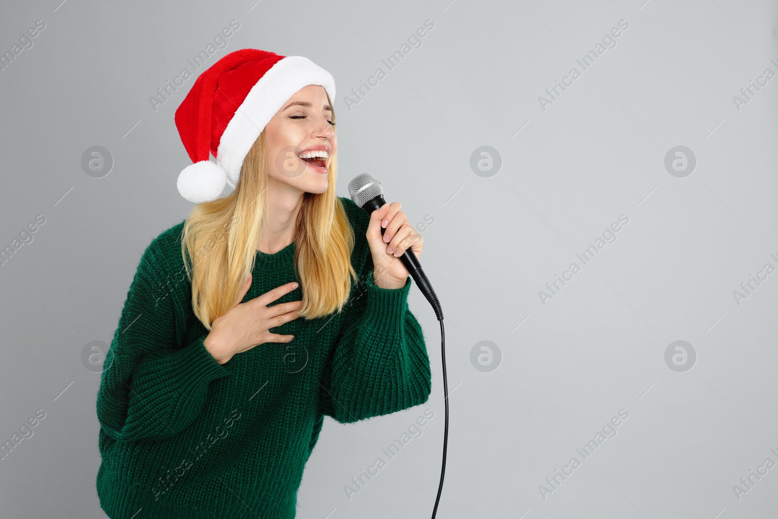 Photo of Emotional woman in Santa Claus hat singing with microphone on grey background, space for text. Christmas music