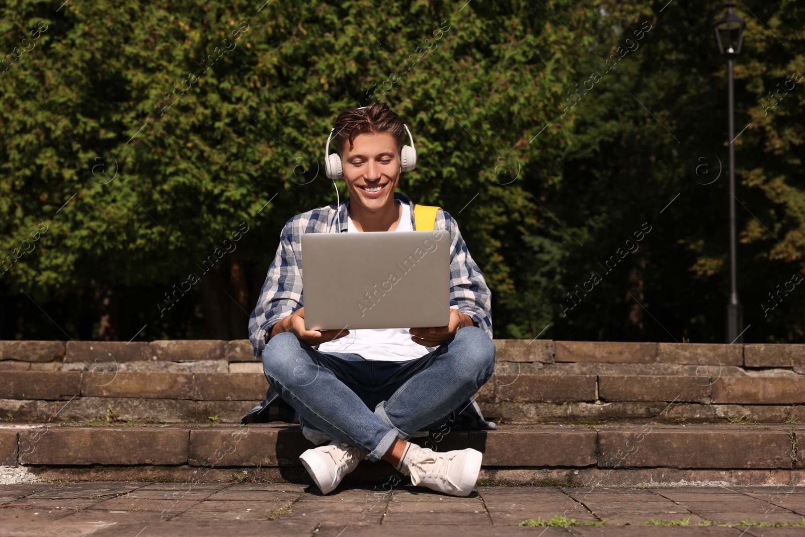 Photo of Happy young student with headphones studying with laptop on steps in park