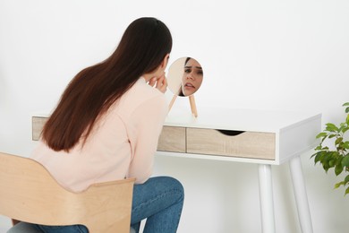 Young woman looking in mirror and touching her face indoors. Hormonal disorders