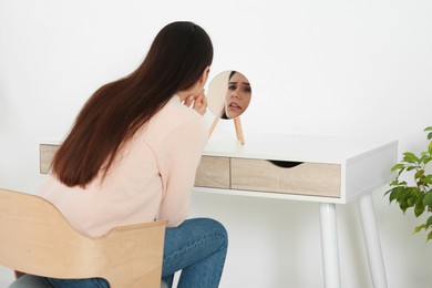 Photo of Young woman looking in mirror and touching her face indoors. Hormonal disorders