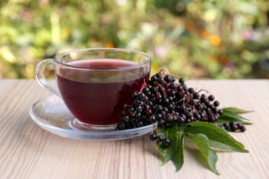 Photo of Glass cup of tasty elderberry tea and Sambucus berries on wooden table outdoors