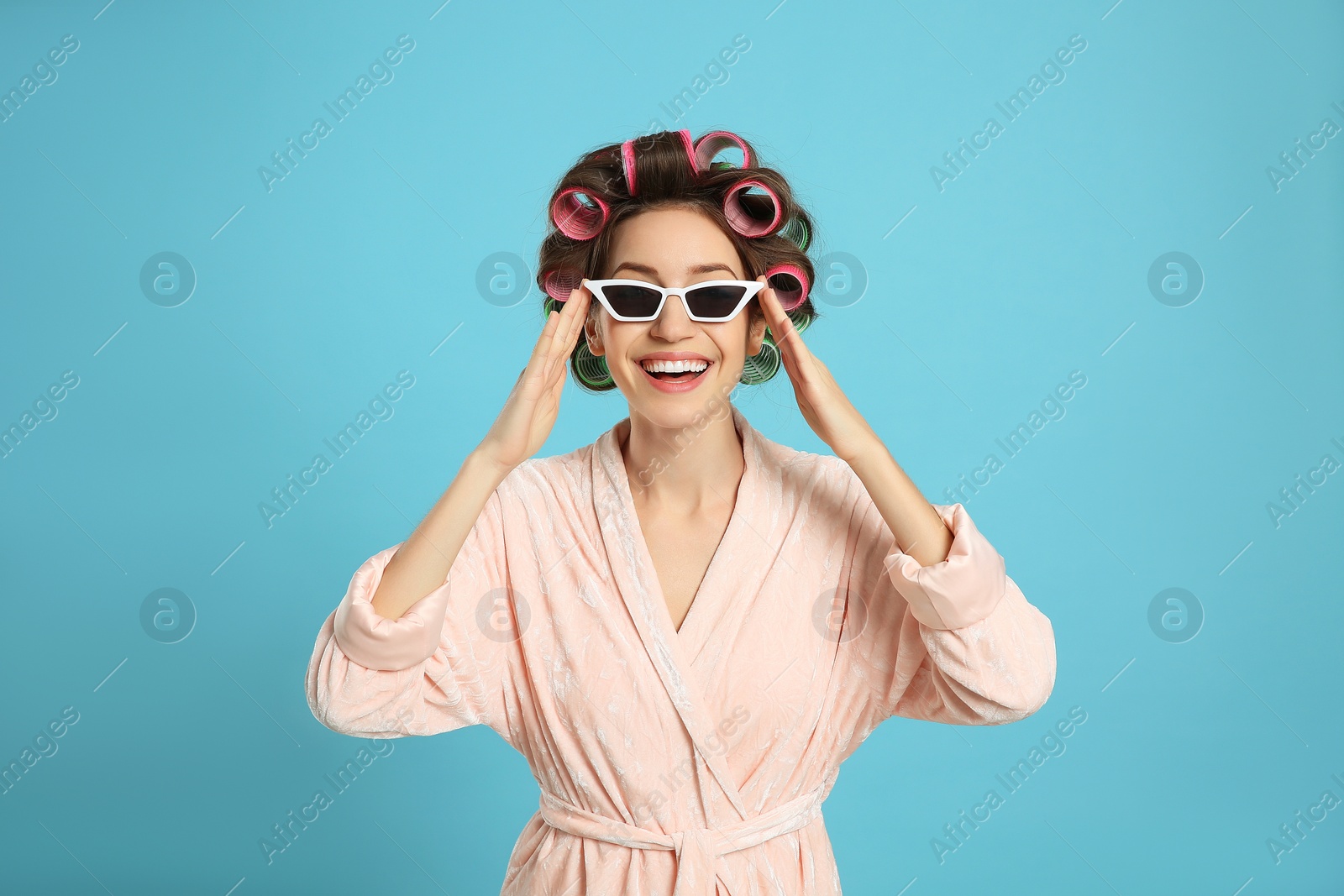 Photo of Beautiful young woman in bathrobe with hair curlers and sunglasses on light blue background