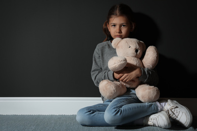 Sad little girl with teddy bear near black wall, space for text. Domestic violence concept