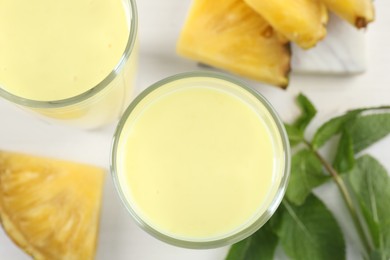 Tasty pineapple smoothie in glasses, mint and cut fruit on white table, flat lay