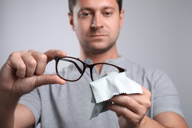 Man wiping glasses with microfiber cloth on light grey background, selective focus