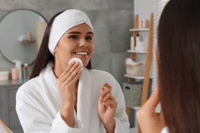 Young woman cleaning her face with cotton pad near mirror in bathroom