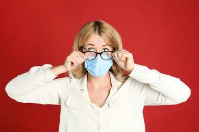 Woman wiping foggy glasses caused by wearing medical mask on red background