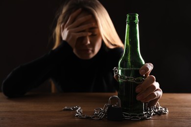Photo of Alcohol addiction. Woman chained with bottle of beer at wooden table against black background, focus on hand