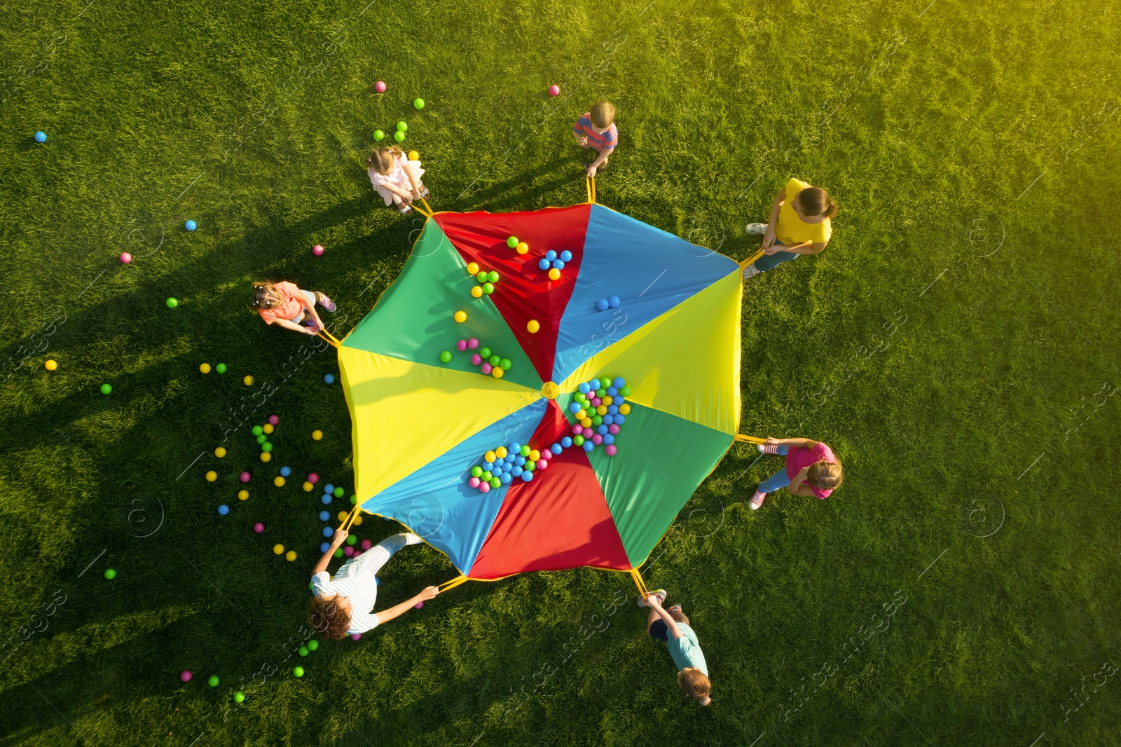 Image of Group of children and teachers playing with rainbow playground parachute on green grass, top view. Summer camp activity