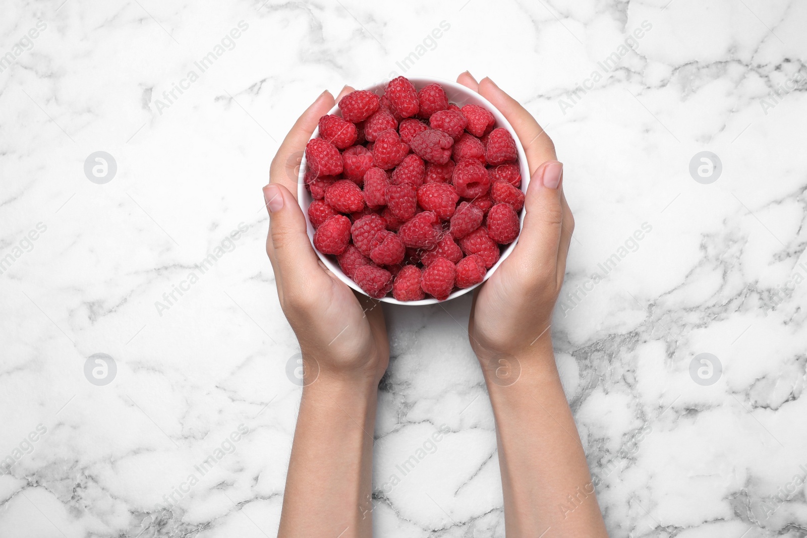 Photo of Woman holding bowl of delicious ripe raspberries at white marble table, top view