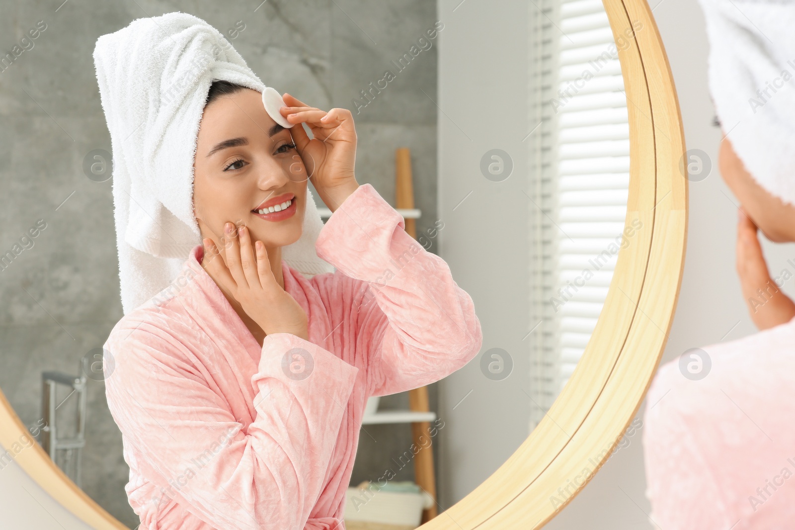 Photo of Beautiful woman in terry towel removing makeup with cotton pad near mirror indoors