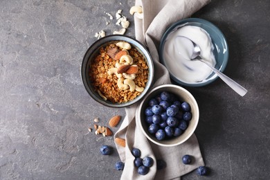 Photo of Tasty granola in bowl, blueberries, yogurt and spoon on gray textured table, flat lay. Space for text