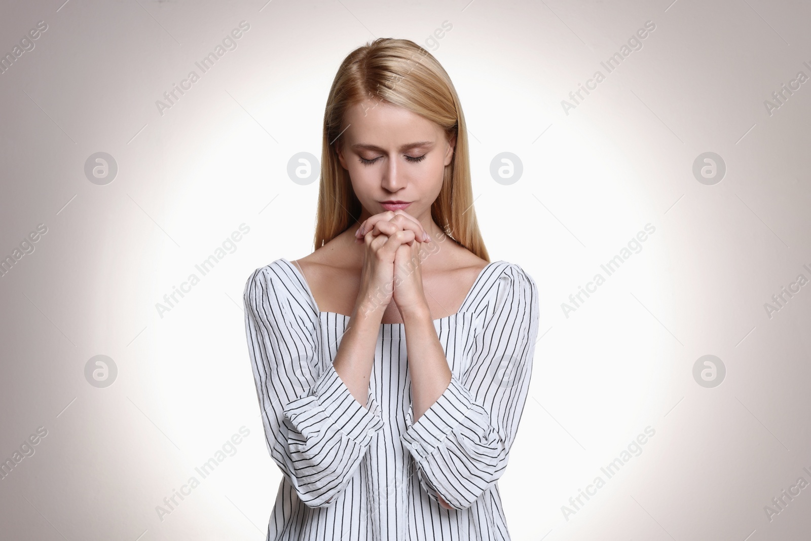 Photo of Religious young woman with clasped hands praying against light background