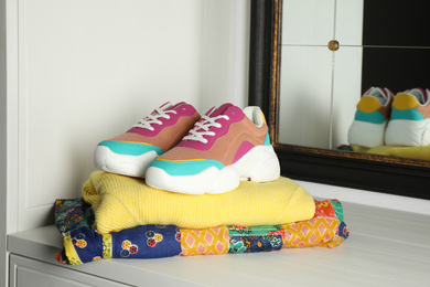 Photo of Stack of folded clothes and colorful sneakers on cabinet indoors