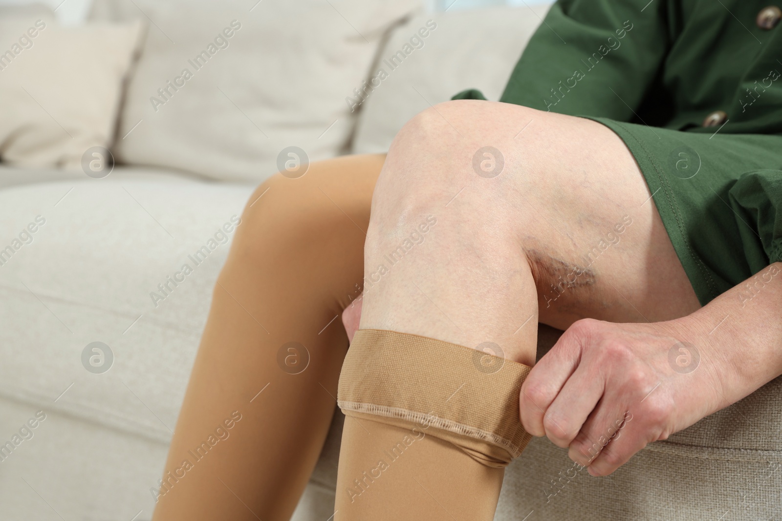 Photo of Woman putting on compression stocking in living room, closeup. Prevention of varicose veins