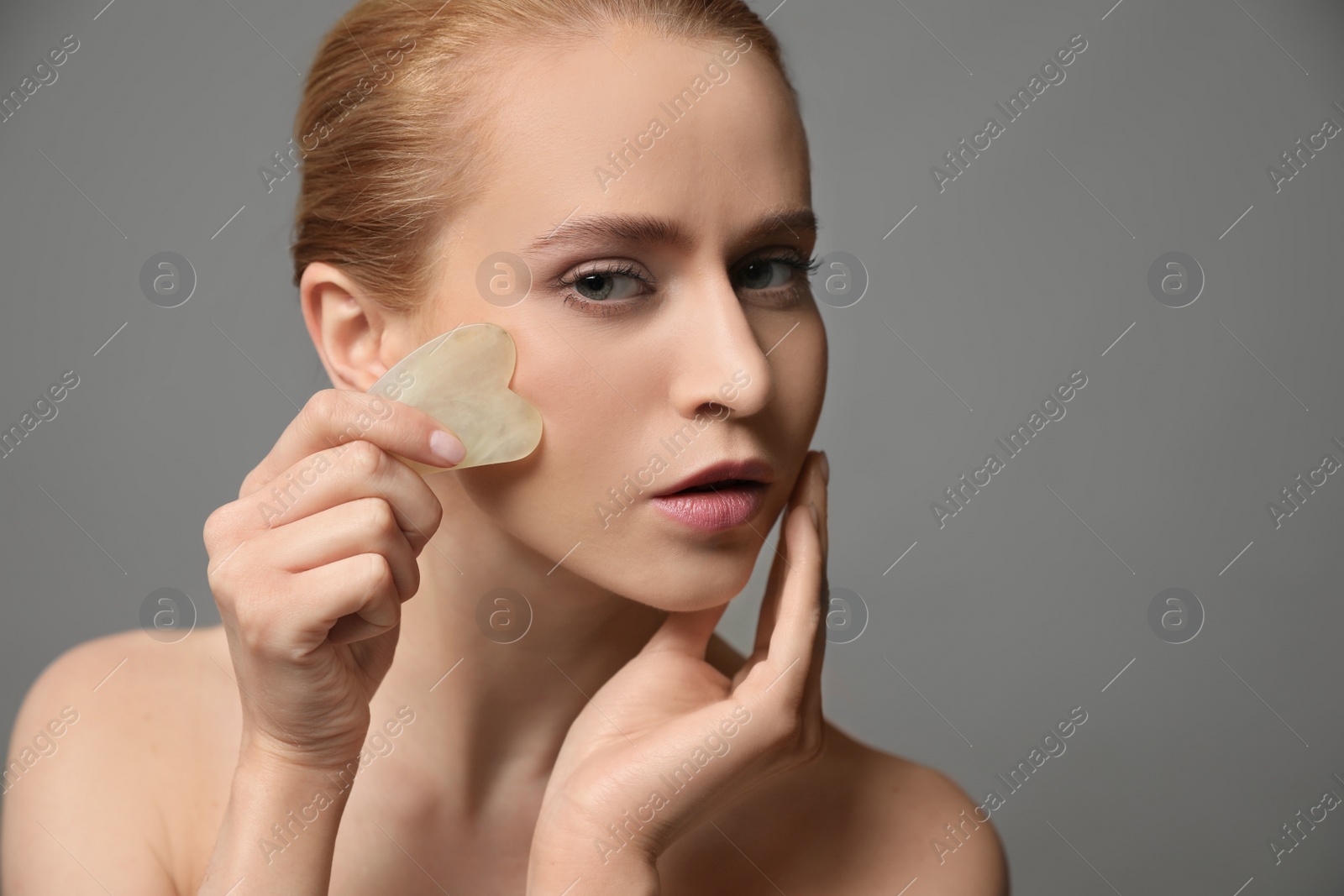 Photo of Beautiful young woman doing facial massage with gua sha tool on grey background, closeup