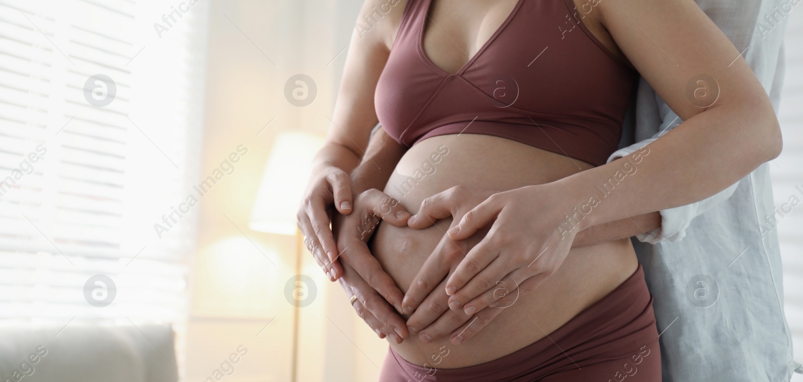 Image of Pregnant young woman making heart with hands on belly and husband near her at home, closeup view with space for text. Banner design