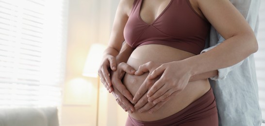Image of Pregnant young woman making heart with hands on belly and husband near her at home, closeup view with space for text. Banner design