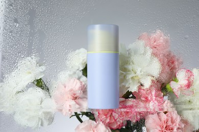 Bottle with moisturizing cream and beautiful flowers on light background, view through wet glass