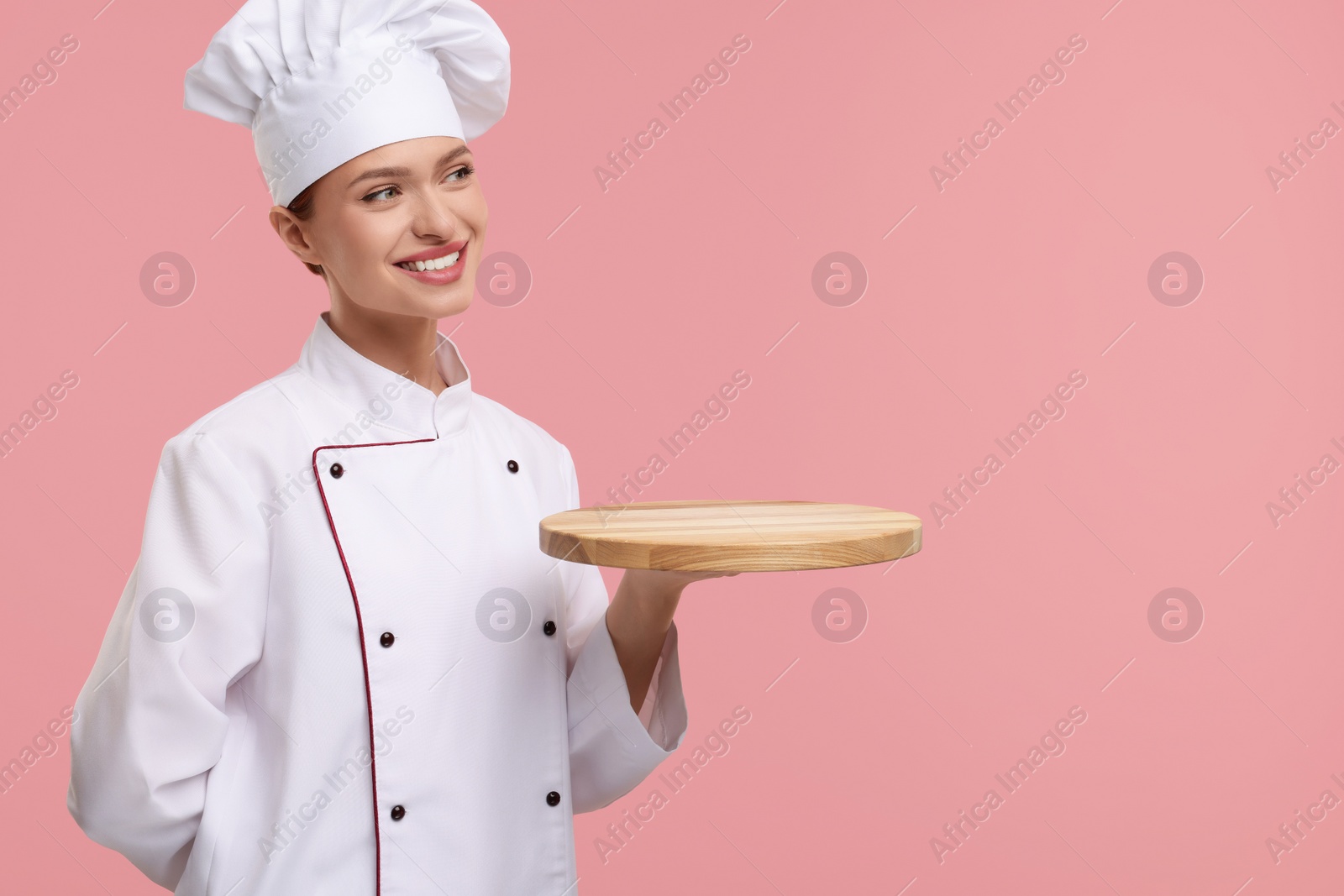 Photo of Happy woman chef in uniform holding empty wooden board on pink background, space for text
