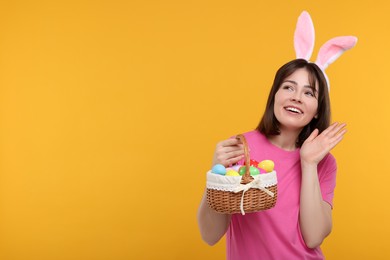 Photo of Easter celebration. Happy woman with bunny ears and wicker basket full of painted eggs on orange background, space for text