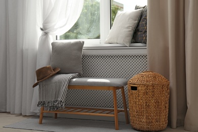 Photo of Striped plaid, hat and cushion on bench near window sill with pillows in room. Cozy place to relax