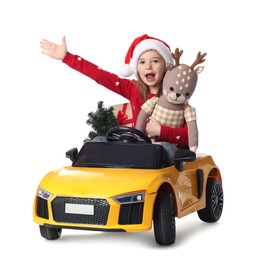 Photo of Cute little girl in Santa hat with Christmas tree and deer toy driving children's car on white background