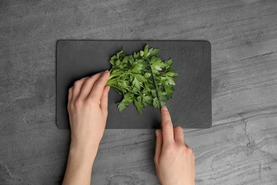 Photo of Woman cutting fresh green parsley on slate board at grey table, top view
