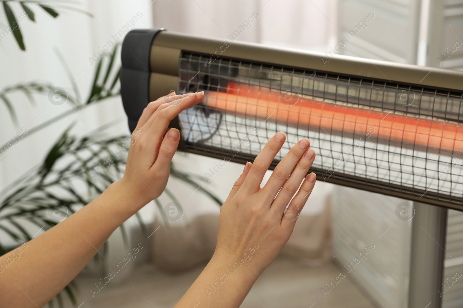 Photo of Woman warming hands near electric infrared heater indoors, closeup