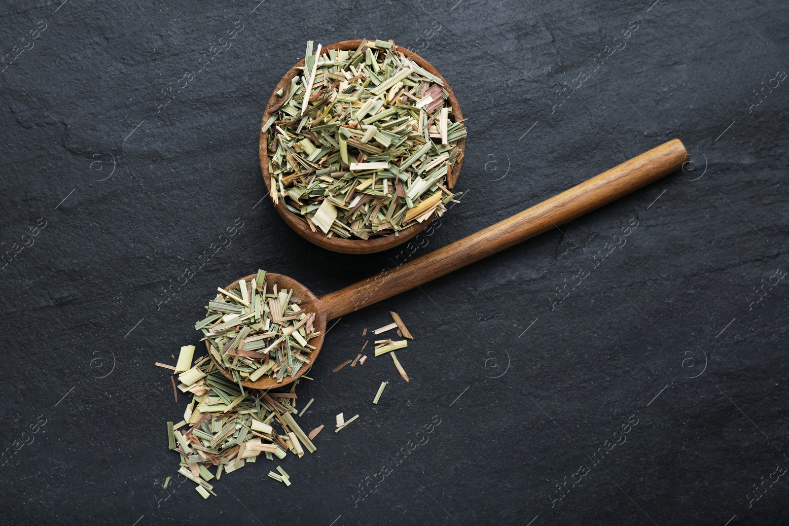 Photo of Wooden bowl and spoon with aromatic dried lemongrass on black table, flat lay