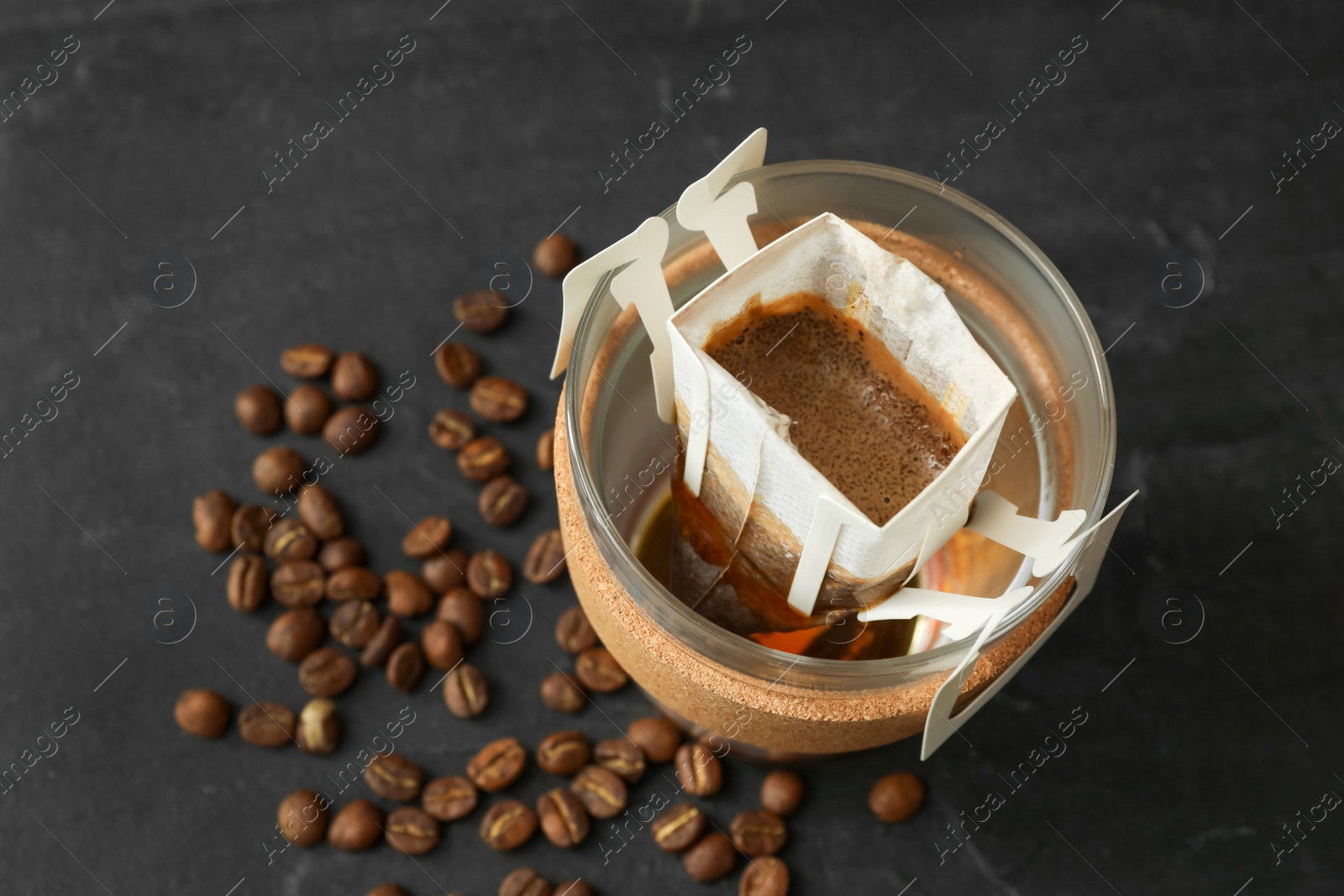 Photo of Glass cup with drip coffee bag and beans on black table, above view